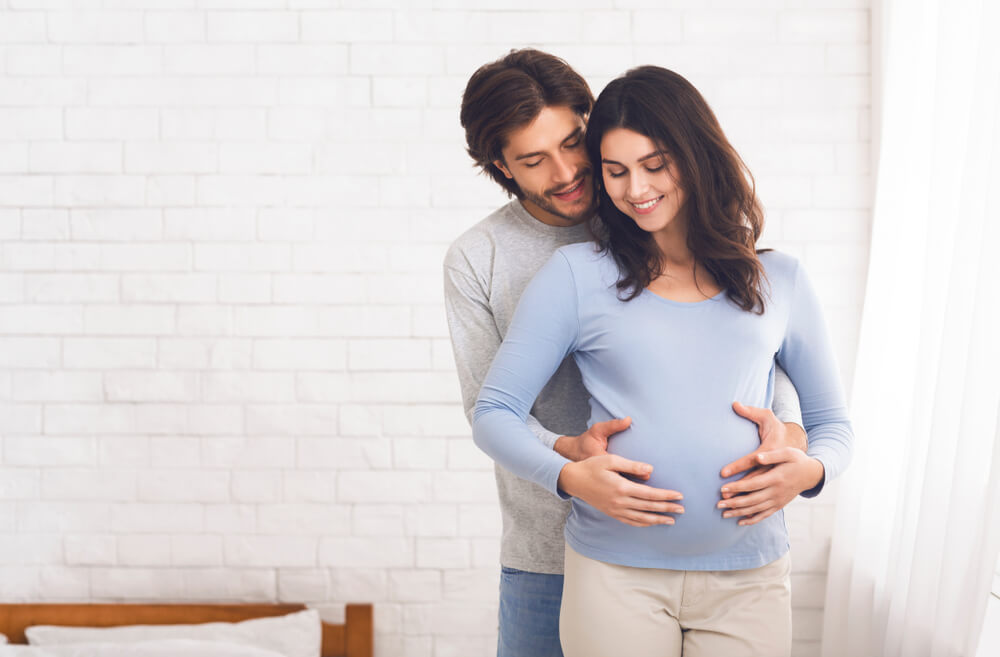 Happy couple expecting baby and hugging near window at home